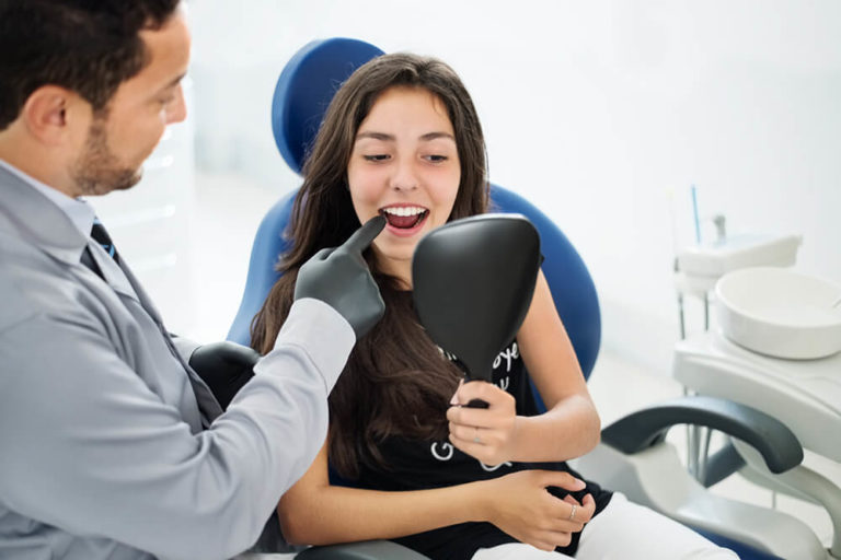 A male dentist points to the teeth of a female patient who is seated in a dental chair and is holding a mirror to see her teeth