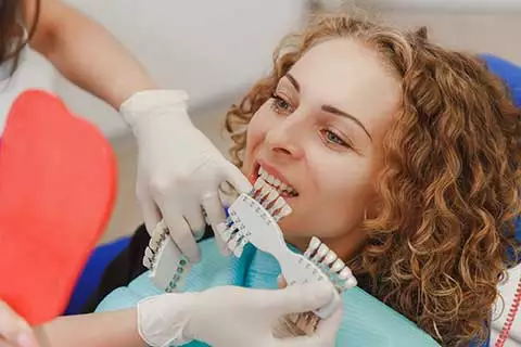 female sites in a dental chair while the dentist compares which shade of tooth color matches the patient's teeth