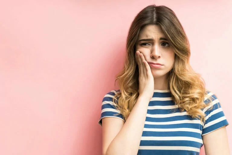 Stock image of a woman holding her cheek in pain while standing against a pink wall.